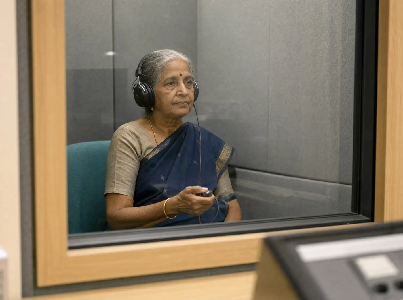 An older woman in the sound treated booth wearing audiometric headphones, holding the response button, seen through the booth window.